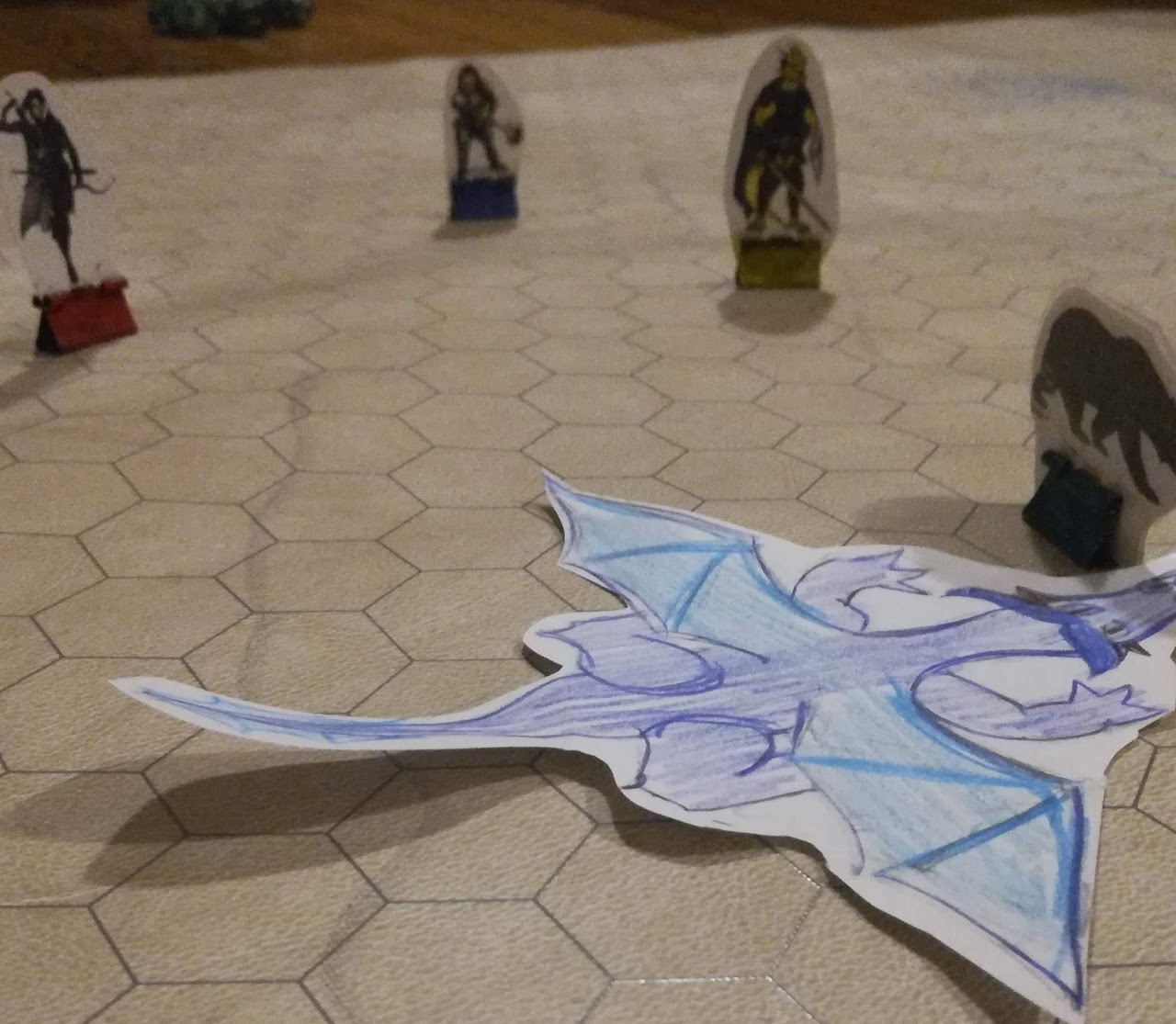 Slanted view of a Dungeons and Dragons map, with several miniatures (including a dwarf, a dragonborn, a bear, and a rogue half-elf) surrounding a paper cutout of a blue dragon.