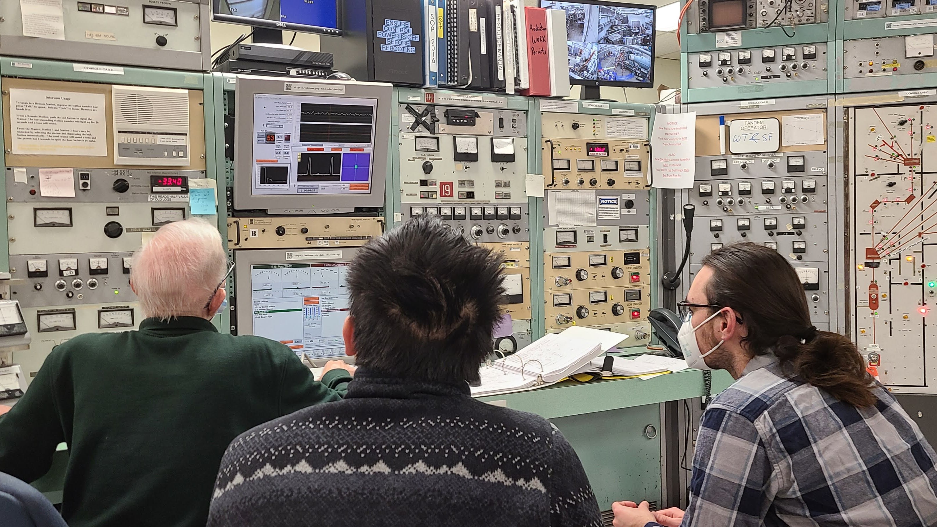 Three researchers sit facing away from the camera in front of a control booth for the TUNL accelerator. The booth is nearly completely covered in dials and knobs, with sticky notes and laboratory books on all empty space. A monitor in the center of the images catches the eyes of the researchers, and shows several pulses and the spread of the neutron beam through a 2-dimensional histogram.