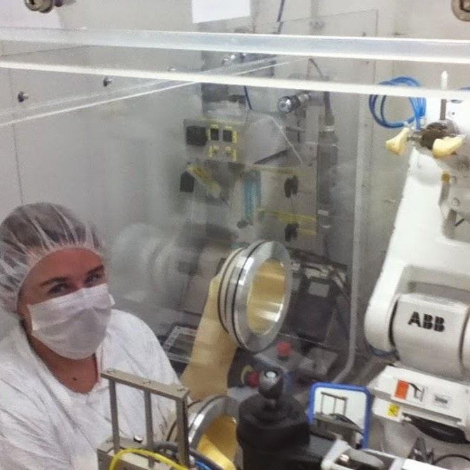 Erin posing at a cleanroom glovebox which contains the CUORE construction robotic arm. The arm has small rubber grips which are prepared to grab a crystal in order to prepare it for processing. Erin is wearing a bunny suit, mask, and hairnet.