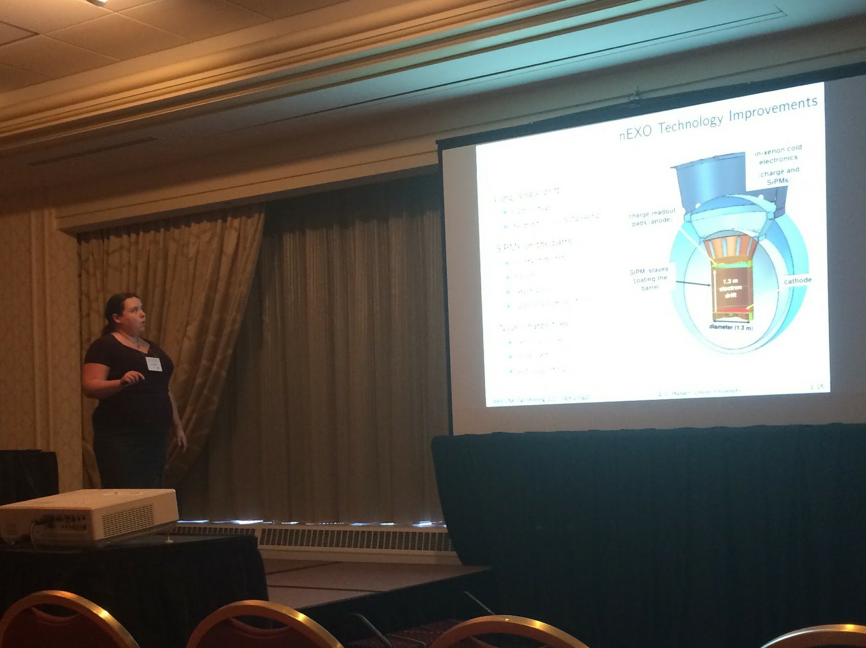 Photograph of Erin giving a talk at a conference. She stands to the left and looks towards the slides projected on a white screen on the right. She uses a laser to point out details on a slide labeled 'nEXO Technology Improvements' which is hard to read but has an image of the nEXO detector in the cryopit at SNO lab